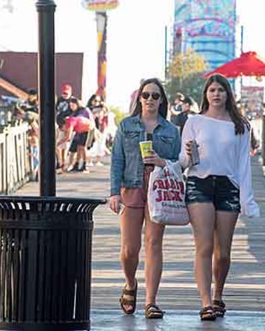 Two women walking on a wooden boardwalk with shops and amusement park rides in background