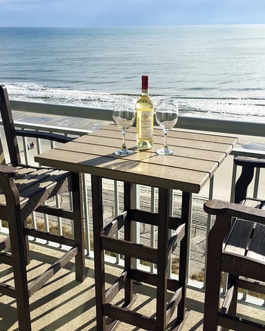 Oceanfront deck with wooden table, chairs, wine bottle and glasses overlooking sandy beach and calm sea