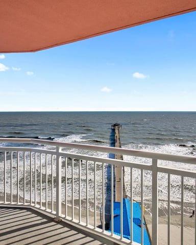 Oceanfront balcony with white railing overlooking sandy beach, pier, and waves under blue sky