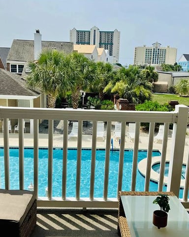 Deck with white railing overlooking swimming pool, palm trees, and beachfront homes with high-rise hotels in background