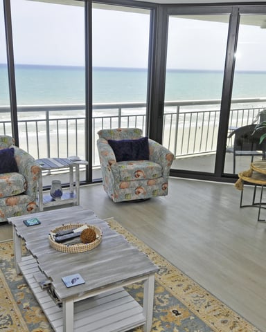 Beachfront living room with patterned armchairs, coffee table, and ocean view through floor-to-ceiling windows and balcony doors