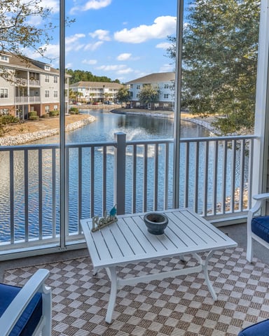 Waterfront deck with blue cushioned seating, small table, and railing overlooking pond and residential buildings