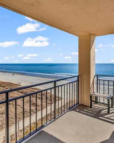 Oceanfront balcony with metal railing overlooking sandy beach, ocean, and blue sky with clouds