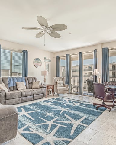 Spacious living room with gray upholstered seating, ceiling fan, blue patterned rug, and glass doors opening to balcony with ocean view.