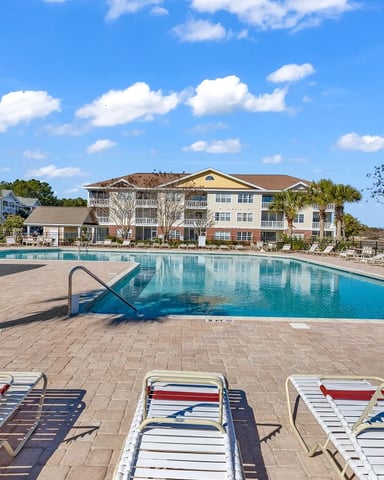 Resort swimming pool with lounge chairs, multi-story apartment building, palm trees, and clear blue sky