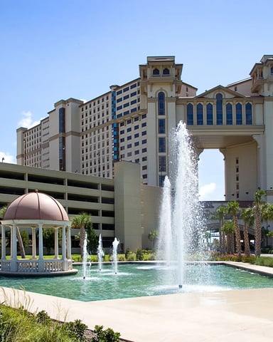 Resort courtyard with fountains, gazebo, palm trees, and large beige buildings with blue-trimmed windows under clear sky