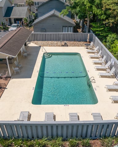 Aerial view of rectangular swimming pool with turquoise water, concrete deck, lounge chairs, and white railing surrounded by residential homes and palm trees.