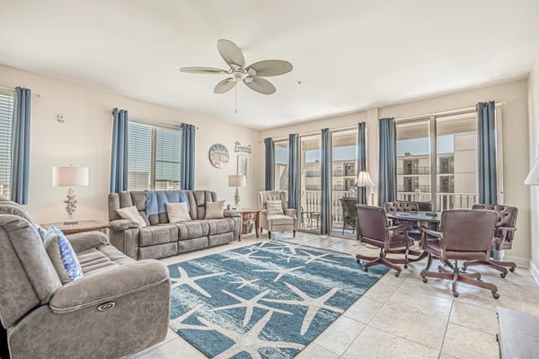 Spacious living room with gray upholstered seating, ceiling fan, blue patterned rug, and glass doors opening to balcony with ocean view.