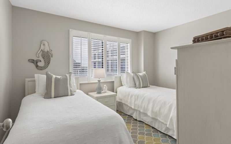 Guest bedroom with two twin beds, white bedding, striped pillows, and shuttered windows with natural light
