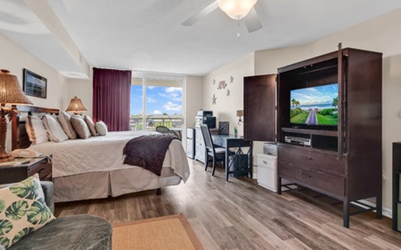 Spacious bedroom with bed, dark wood entertainment center with TV, ceiling fan, and mountain view through windows