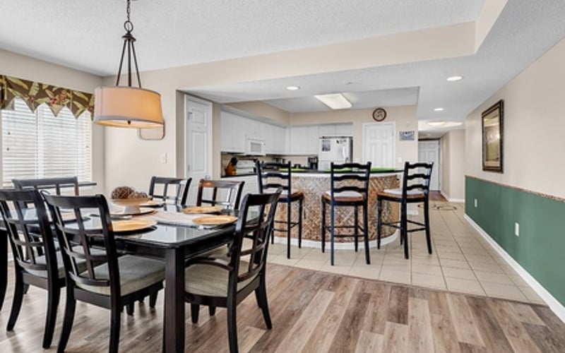 Dining room with black table and chairs, open kitchen visible beyond, pendant light overhead, wood-look flooring