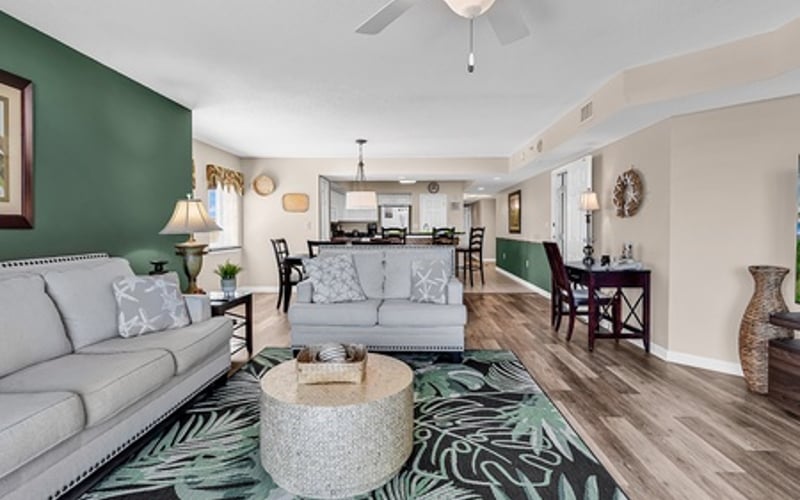 Open-plan living room with gray sofa, green accent wall, ceiling fan, and tropical patterned rug leading to kitchen