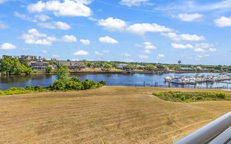Waterfront lawn with marina, river, and residential buildings under blue sky with white clouds