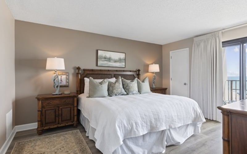 Master bedroom with taupe walls, white bedding, wood nightstands with lamps, and balcony access