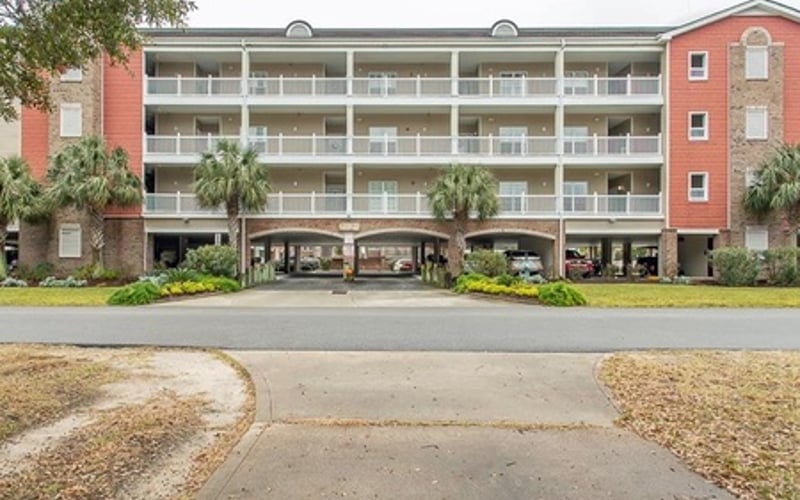 Three-story brick apartment building with covered ground-floor parking and palm trees, driveway approach