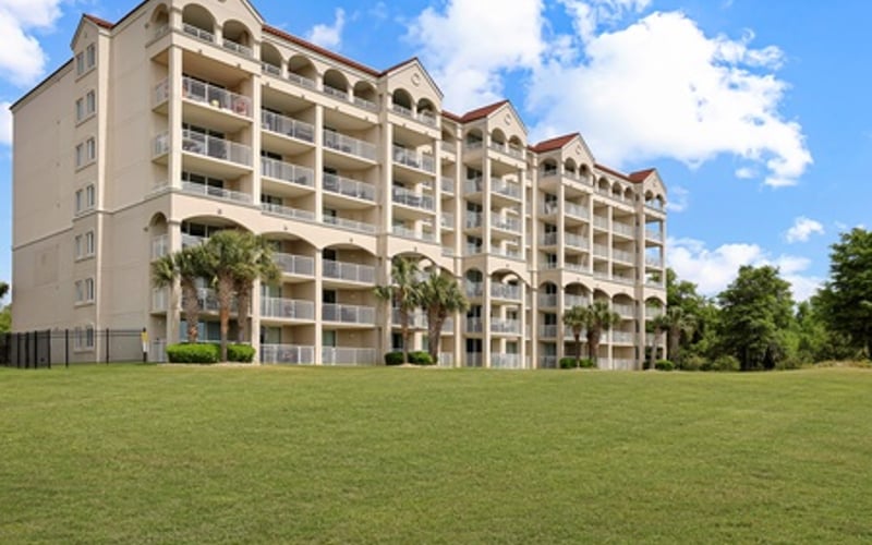 Multi-story beige residential building with balconies and palm trees, viewed from green lawn