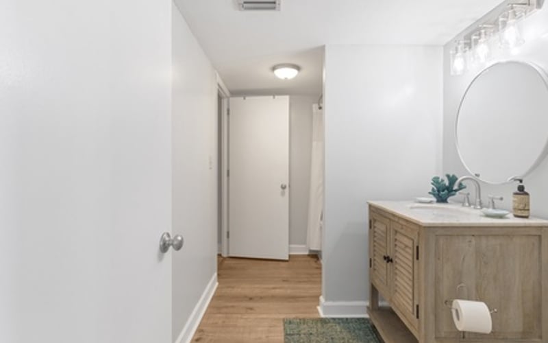 Modern bathroom with wooden vanity, round mirror, and hardwood flooring leading to hallway
