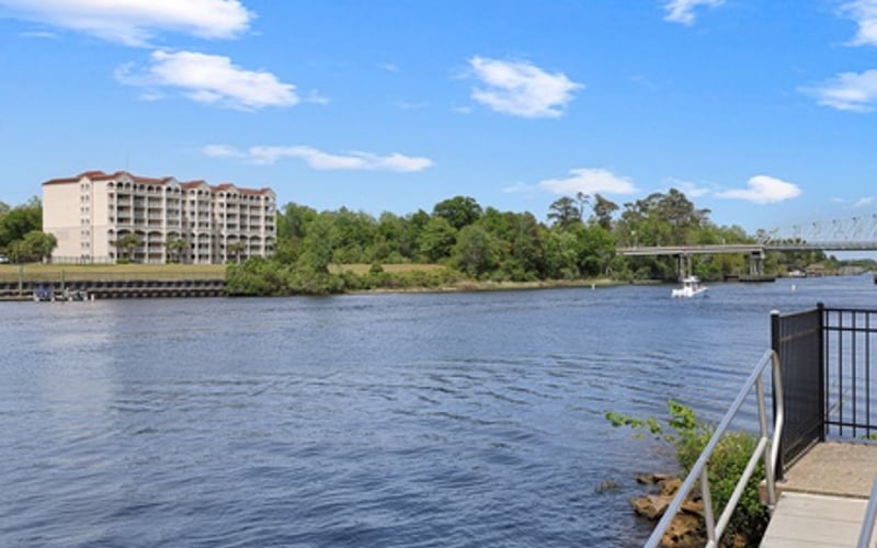 Waterfront view with multi-story residential building, river, sailboat, and bridge under blue sky