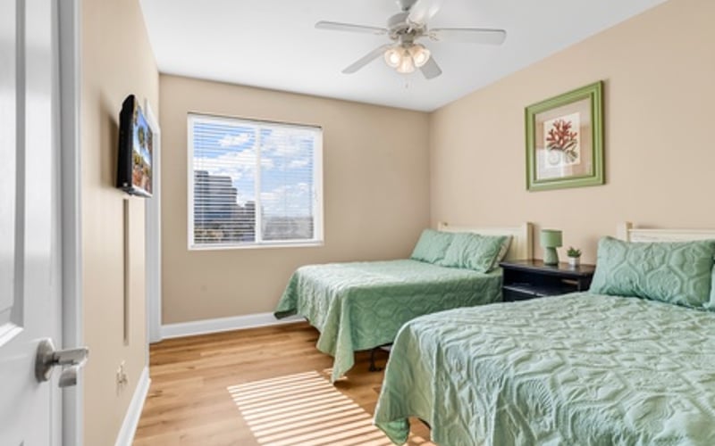 Bedroom with two twin beds with green bedding, ceiling fan, window with blinds, and hardwood flooring