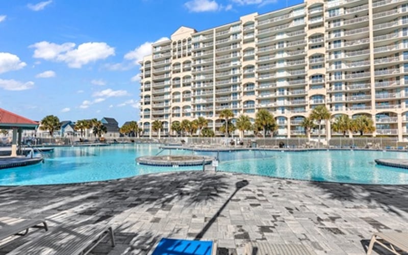 Multi-story beachfront resort building with turquoise pool, lounge chairs, and palm trees under blue sky