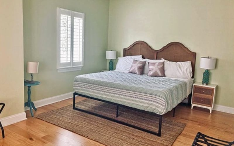 Bedroom with wooden bed frame, white bedding, green accent wall, hardwood floor, and window with shutters