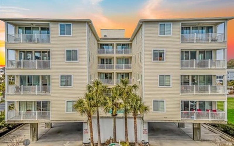 Four-story beige apartment building with white railings and balconies, palm trees in front, sunset sky