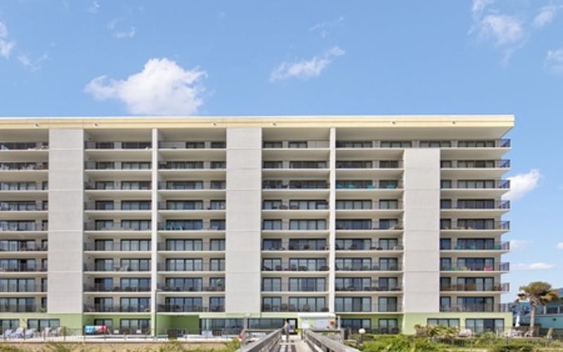 Modern multi-story beachfront apartment building with white facade and balconies, viewed from wooden boardwalk