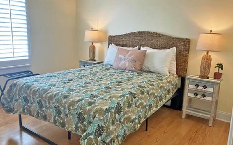 Bedroom with woven headboard, patterned bedspread, nightstands with lamps, and wooden flooring