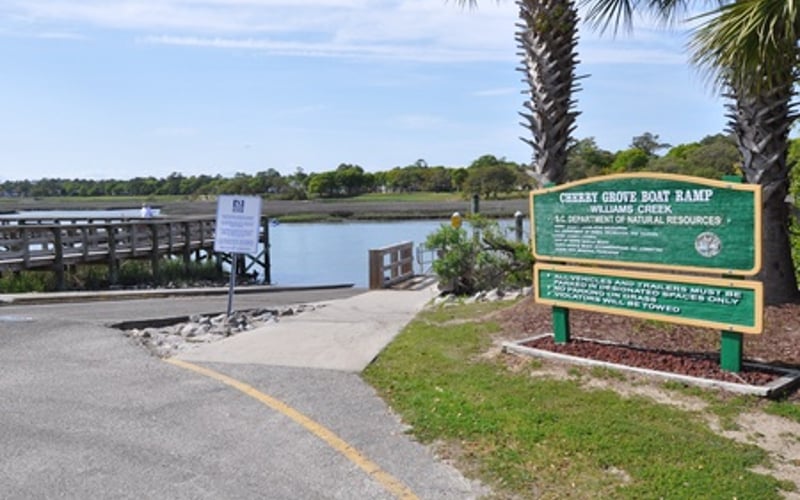 Waterfront park entrance with green informational sign, palm trees, and wooden dock overlooking calm water