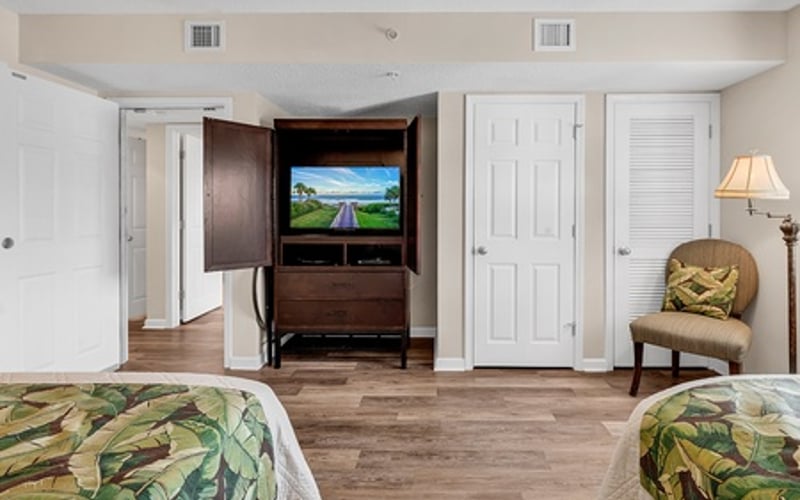 Bedroom with two beds, wooden TV cabinet with landscape display, white closet doors, and upholstered chair with lamp
