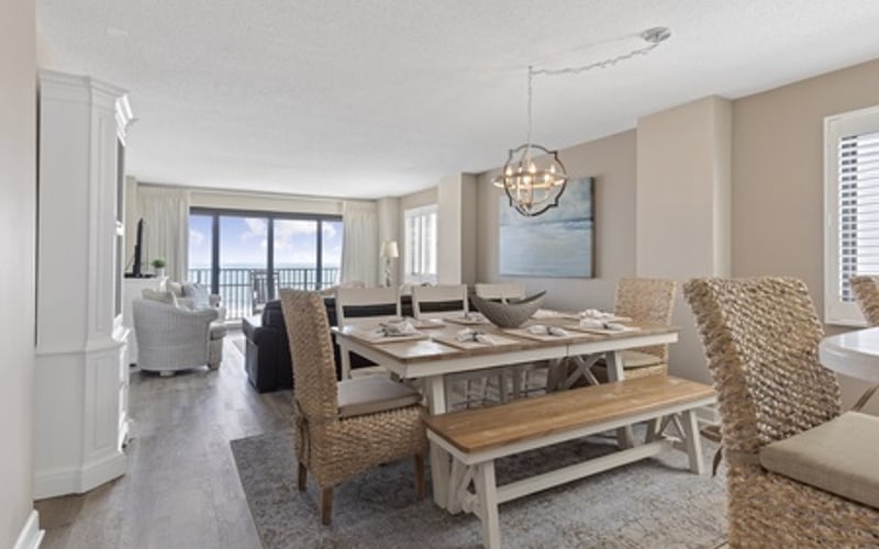 Open-plan dining and living area with wooden table, woven chairs, pendant light, and ocean view through glass doors