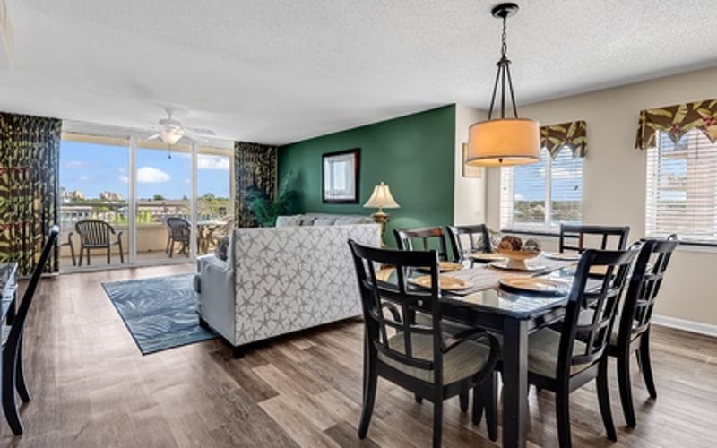Open-plan living and dining area with green accent wall, patterned sofa, dark wood dining table, and ocean view through glass doors