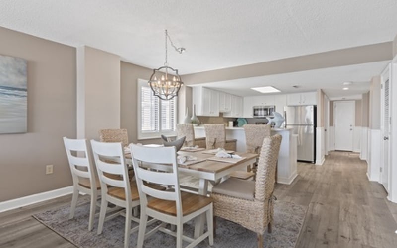 Open-plan dining and kitchen area with white chairs, wooden table, pendant light, and beige walls