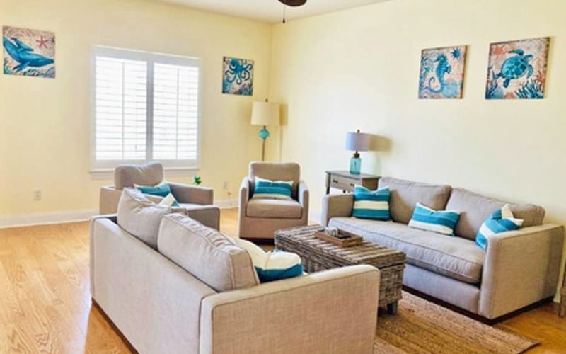 Bright living room with beige sofas, wooden coffee table, ceiling fan, and blue coastal artwork on yellow walls