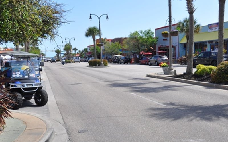 Sunny street with palm trees, parked cars, storefronts, and street lamps