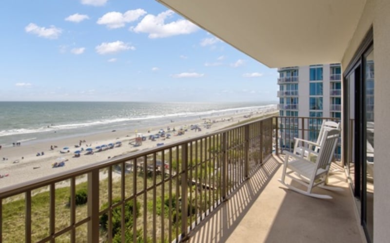 Oceanfront balcony with railing overlooking sandy beach, ocean, and beachgoers on sunny day