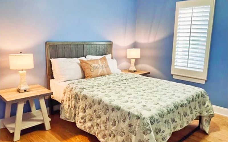 Bedroom with blue walls, wooden bed frame, patterned quilt, nightstands with lamps, and white shutters