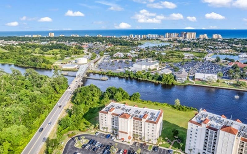 Aerial view of waterfront residential buildings with parking lot, river, and coastal city skyline in distance