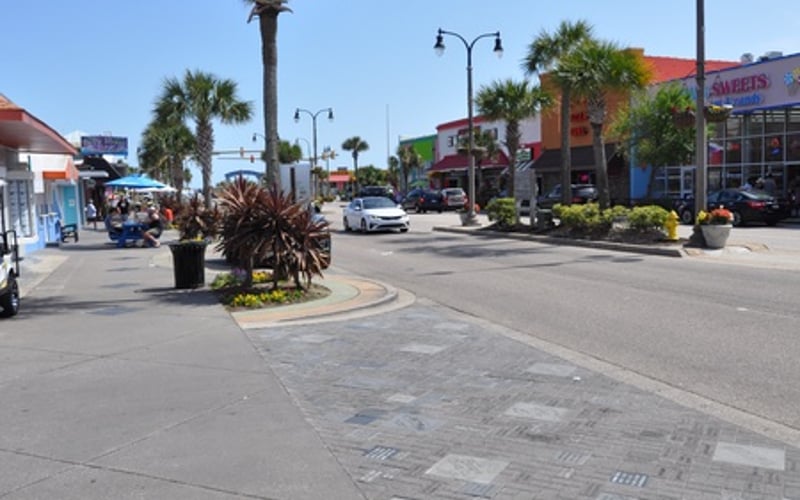 Sunny street with palm trees, storefronts, and parked cars in a coastal commercial area