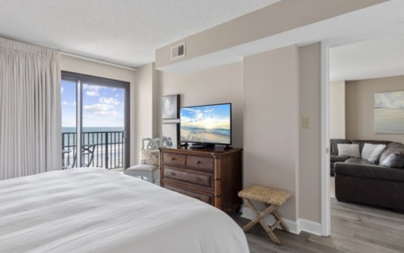 Bright bedroom with white bedding, wooden dresser with TV, balcony doors overlooking water, and open doorway to living area