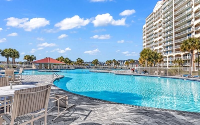 Resort pool with lounge chairs, high-rise building, palm trees, and clear blue sky