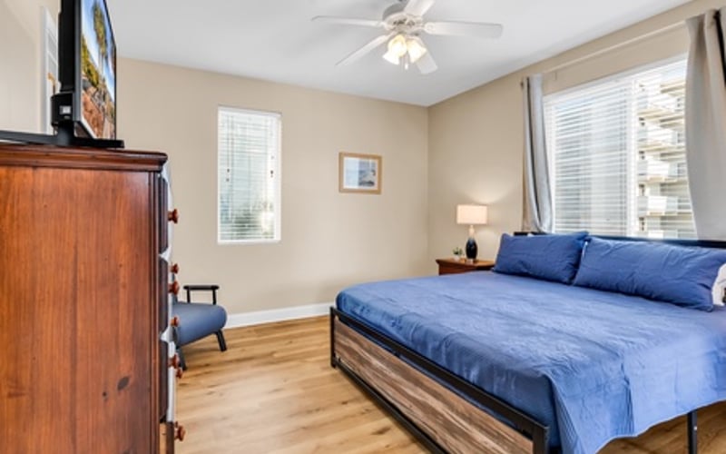 Bedroom with blue bedding, wooden dresser, TV, ceiling fan, and windows with blinds