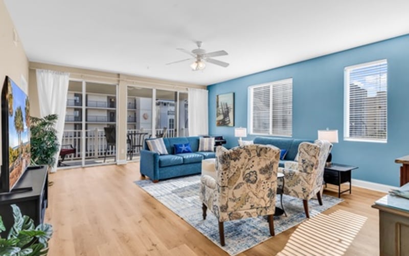 Bright open-plan living room with blue walls, blue sofa, patterned armchairs, wood flooring, and glass doors opening to balcony