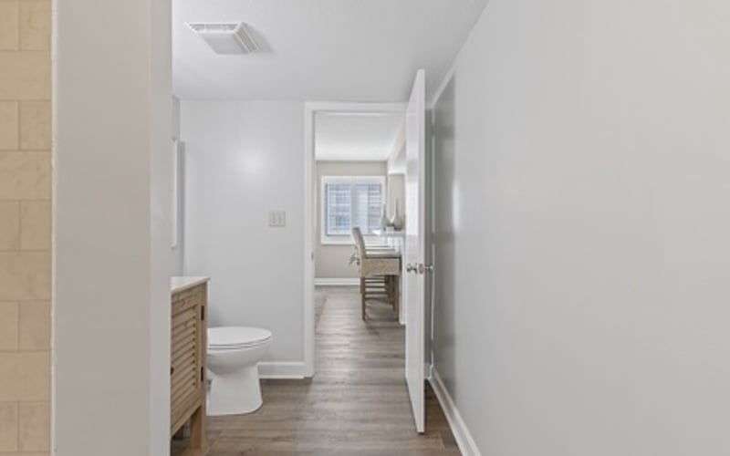 Narrow bathroom hallway with toilet, wooden vanity, and view into bedroom beyond