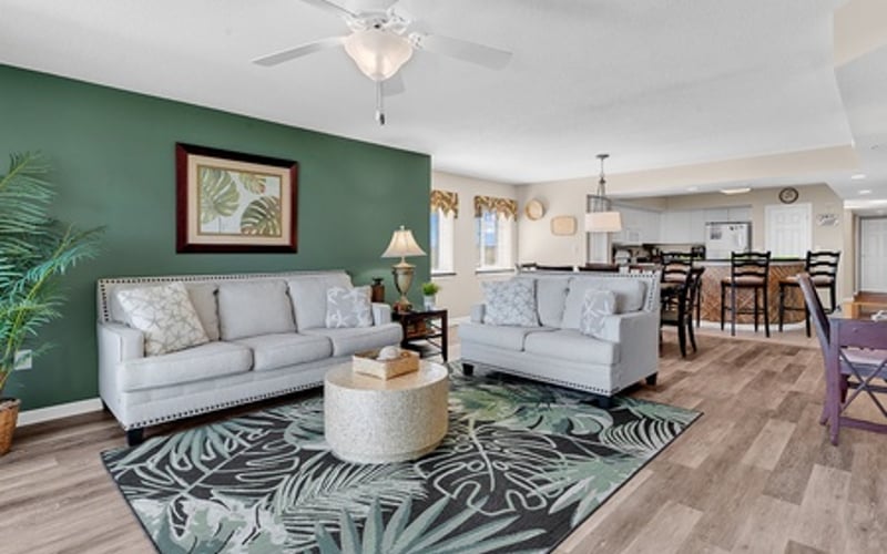 Open-plan living room with two gray sofas, green accent wall, ceiling fan, and tropical patterned rug