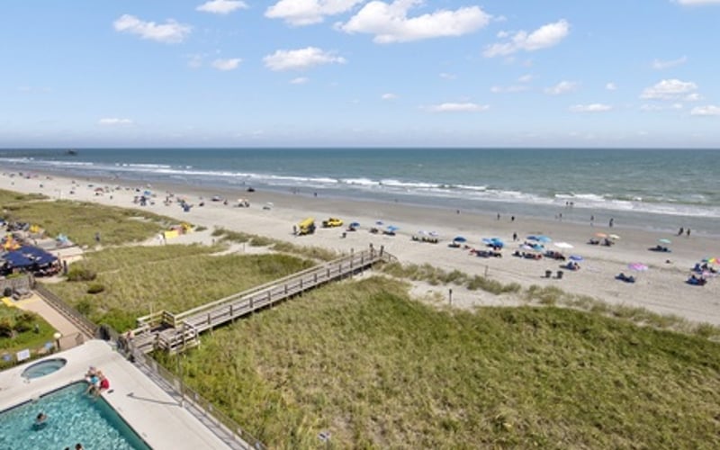 Aerial view of beachfront property with swimming pool, wooden boardwalk through dunes, and sandy beach with beachgoers and vehicles
