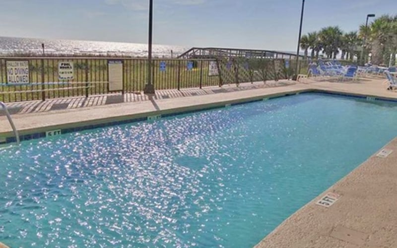 Rectangular swimming pool with turquoise water, metal railing, palm trees, and beach in background