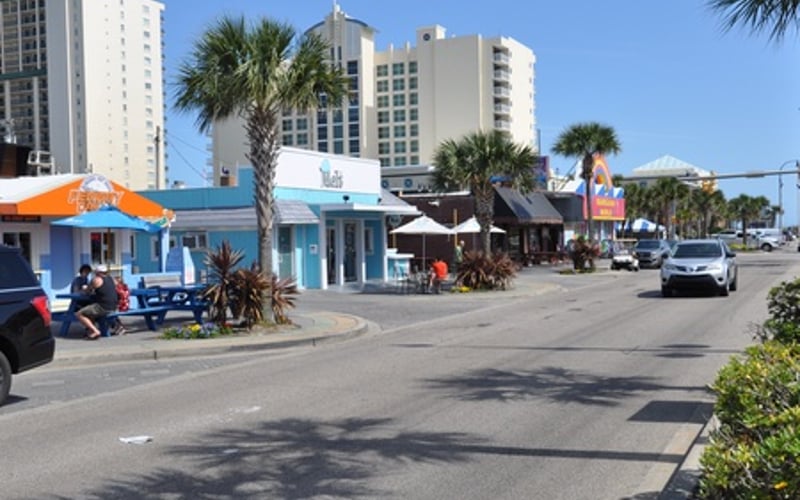 Beachfront commercial street with palm trees, multi-story hotels, and shops under clear blue sky