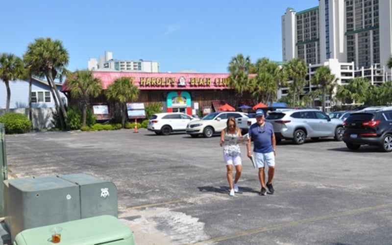 Beachside parking lot with vintage arcade building, parked cars, and two people walking toward the street
