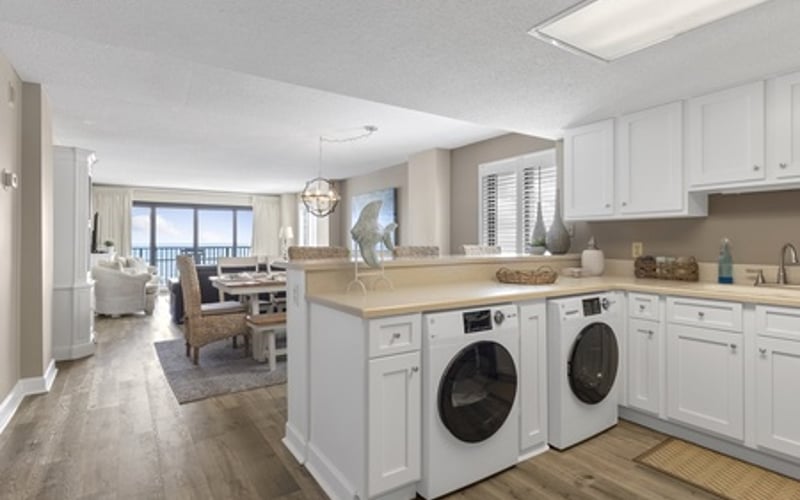 Modern laundry room with white cabinetry, two front-load washers, and wood countertops, opening to living area with ocean view